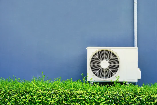 Scène extérieure avec unité de climatisation blanche installée sur support métallique contre mur bleu, entourée de végétation verte dense