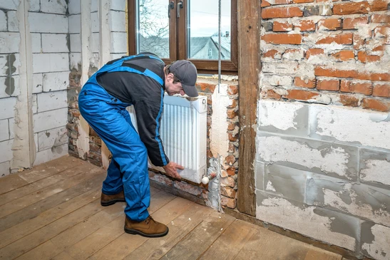 Ouvrier en salopette bleue installant un radiateur blanc contre un mur en briques apparentes dans une maison en rénovation